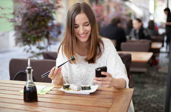 Счастливый молодая женщина ест суши в ресторане и с помощью мобильных — стоковое фото