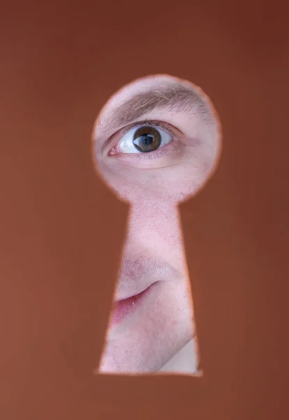 Человек глаз смотрит через отверстие в замочную скважину, на коричневый фон — стоковое фото