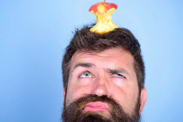 Hipster unhappy face with apple stump target on head blue background, close up. Man handsome hipster long beard almost eaten apple stump on head as target. Dieting goal. Live target concept — стоковое фото