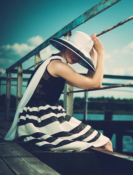 Красавица, носящая шляпу и белый шарф, сидящий на старом деревянном пирсе — стоковое фото