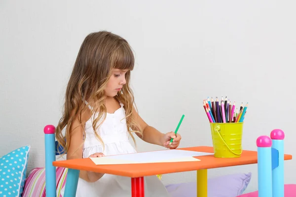 Маленькая девочка рисует сидя за столом в комнате на фоне серой стене — стоковое фото