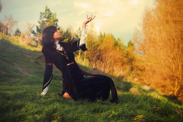 Красивая молодая женщина с темными волосами и историческое платье позирует на лугу в открытом пейзаж с жестом связи между небесной и земля миры — стоковое фото