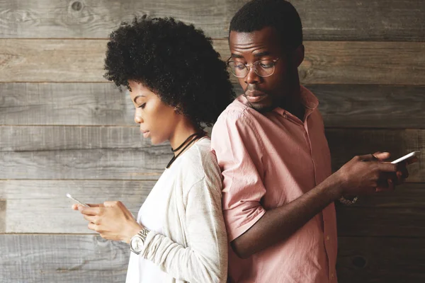 Black girl with Afro haircut texting her lover on cell phone while her jealous boyfriend, suspect of her betrayal, trying to read what she is typing, looking over his shoulder with curious expression — стоковое фото