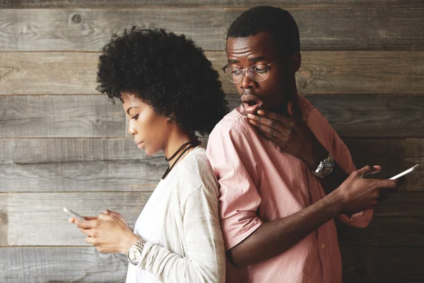 Surprised concerned African man looking at the screen of his girlfriend's mobile phone with mouth wide open and a hand at his mouth while she is cheating on him texting her lover via social networks — стоковое фото