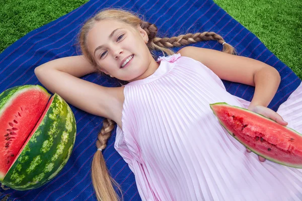 Милая маленькая девочка ест арбуз на траве в летнее время. с длинными волосами, хвост и зубастая улыбка сидя на траве и наслаждаясь — стоковое фото