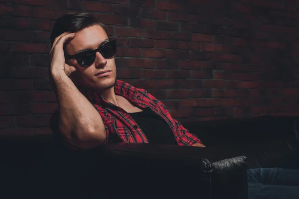 Портрет битник красивый молодой человек очки повседневную одежду расстегнутую рубашку сидя расслабляющий, опираясь на кожаный диван люкс смотрит в камеру Стоковое Фото