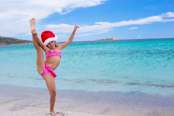 Улыбается маленькая девочка, делая растяжку упражнение в колпак Санта-Клауса на белом пляже — стоковое фото