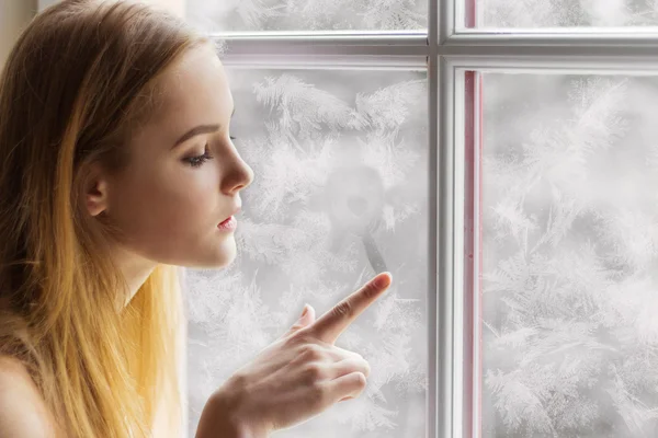 Красивая молодая девушка, сидящая у дня зимы окна и, тянет солнце на замороженном окне — стоковое фото