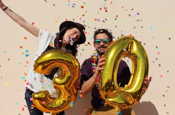 Веселая пара празднует день рождения 30 лет с большой золотые шары и красочные маленькие кусочки бумаги в воздухе — стоковое фото