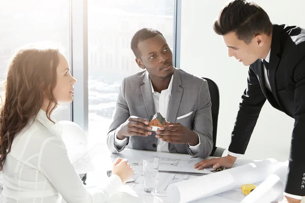 African boss holding scale model house of future real estate while two young Caucasian engineers presenting their architectural project to him. Group of architects discussing business plans in office — стоковое фото