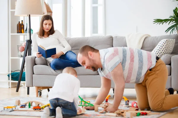 Фото семьи отца и сына, играя на полу в дороге игрушку на фоне беременной женщины, читать книги, лежа на диване — стоковое фото