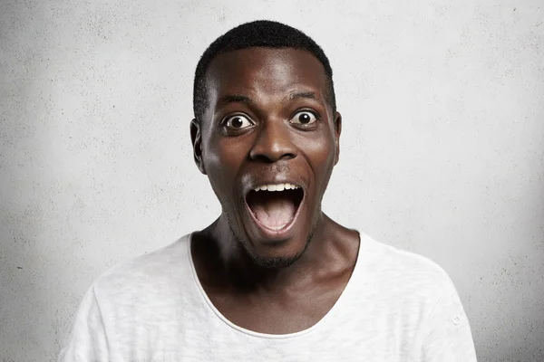 Omg! Portrait of astonished wide-eyed African customer in white t-shirt looking at camera in surprise and astonishment, shocked with unprecedentedly low prices, screaming with mouth wide open — стоковое фото