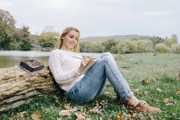 Pretty smiling young woman in white sweater and blue jeans draws in a notebook with colored pencils sitting on the grass and leaned into the trunk of a dry tree on a background of lake — стоковое фото