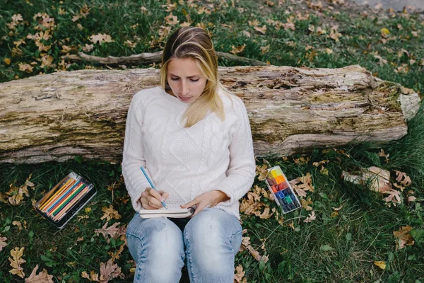 Pretty young woman in white sweater and blue jeans draws in a notebook with colored pencils sitting on the grass and leaned into the trunk of a dry tree — стоковое фото