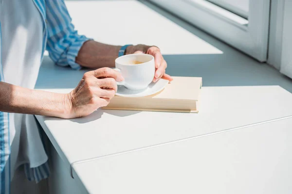 Partial View Senior Woman Book Cup Coffee Windowsill Home — стоковое фото