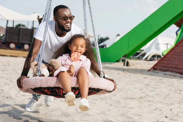 African American Father Pushing Daughter Spider Web Nest Swing Lollipop — стоковое фото