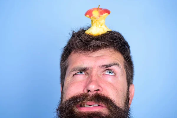 Man handsome hipster long beard almost eaten apple stump on head as target. Dieting goal. Live target concept. Hipster unhappy face with apple stump target on head blue background, close up — стоковое фото